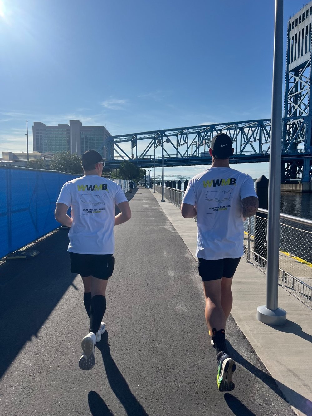 Two runners in Walk With Me, Brother shirts running together beside a bridge.